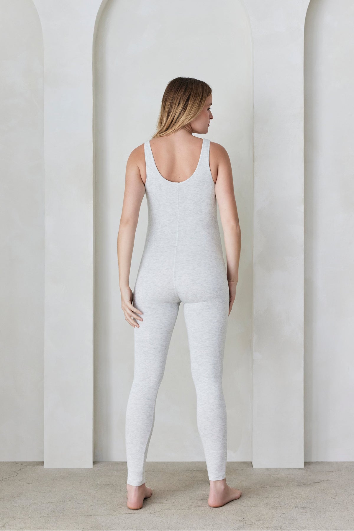 Back view of a woman wearing a heather grey Bumpsuit standing in front of a minimalist background with arches.