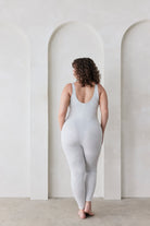 Woman wearing a grey bodysuit standing in a minimalistic room with white walls and arches.