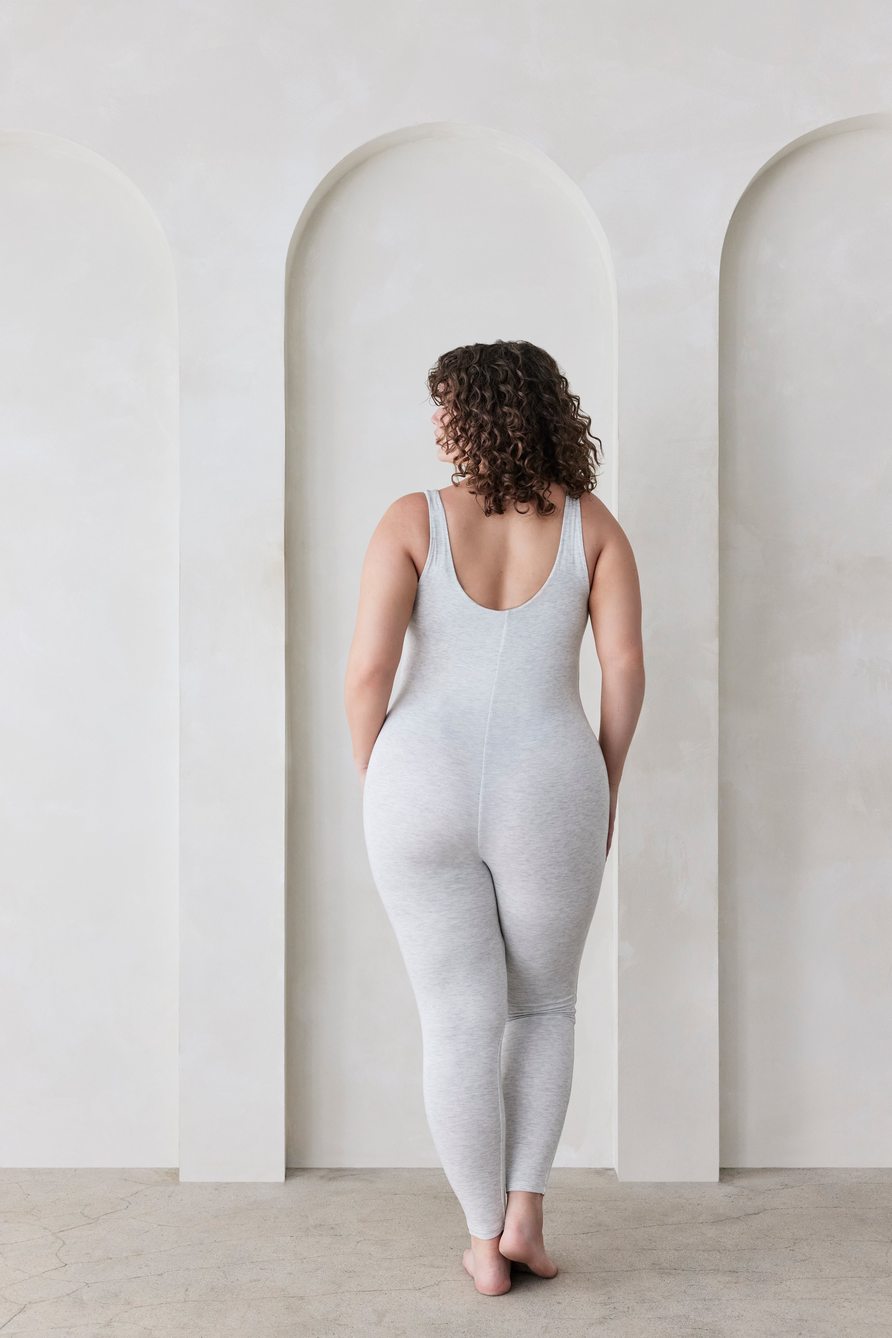 Woman wearing a grey bodysuit standing in a minimalistic room with white walls and arches.