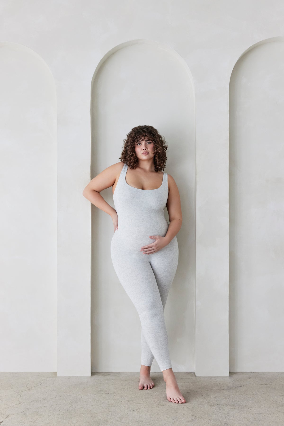 Woman wearing a grey bodysuit standing against a light-colored wall with arches.