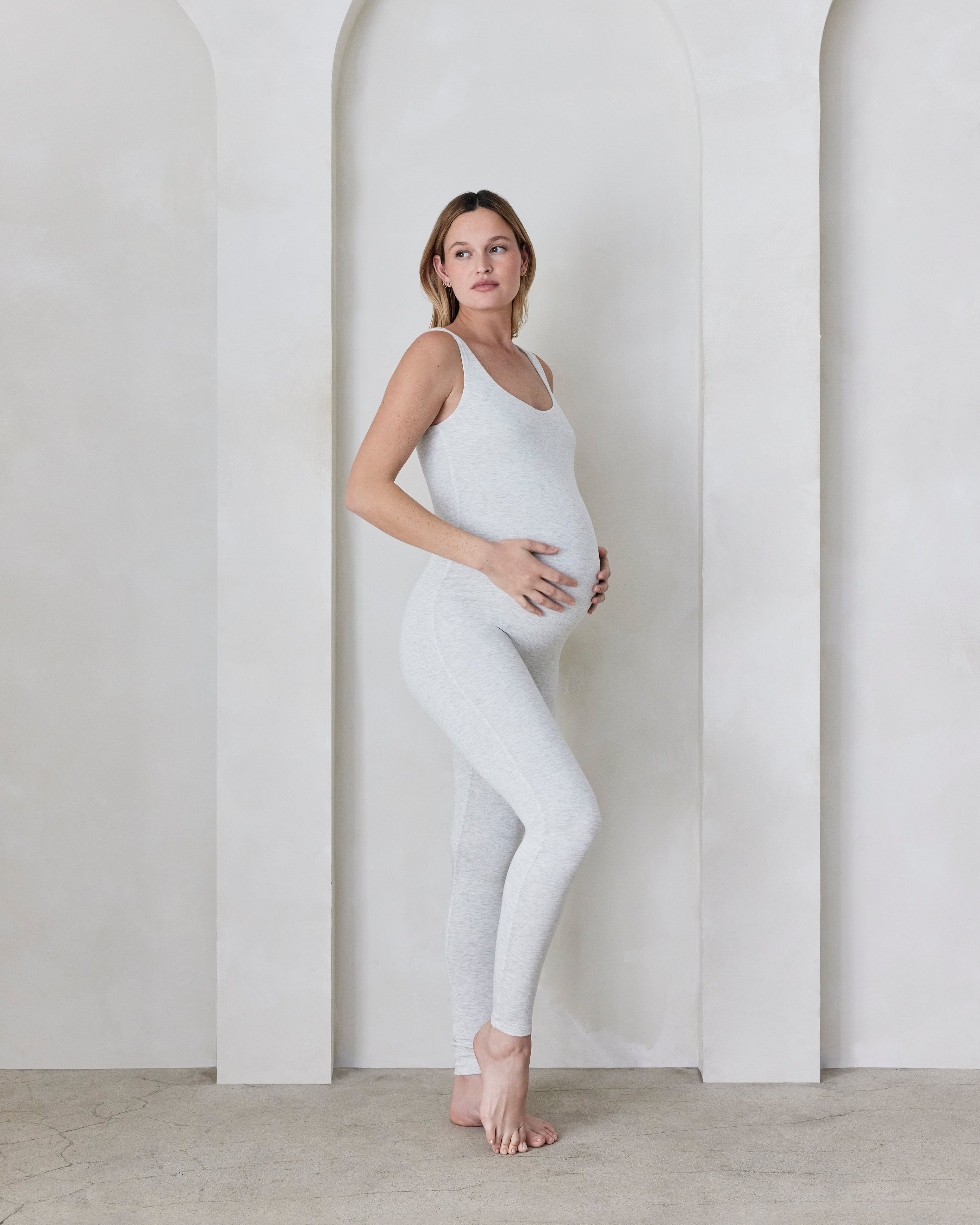 Pregnant woman in a light grey bodysuit standing against a minimalistic background