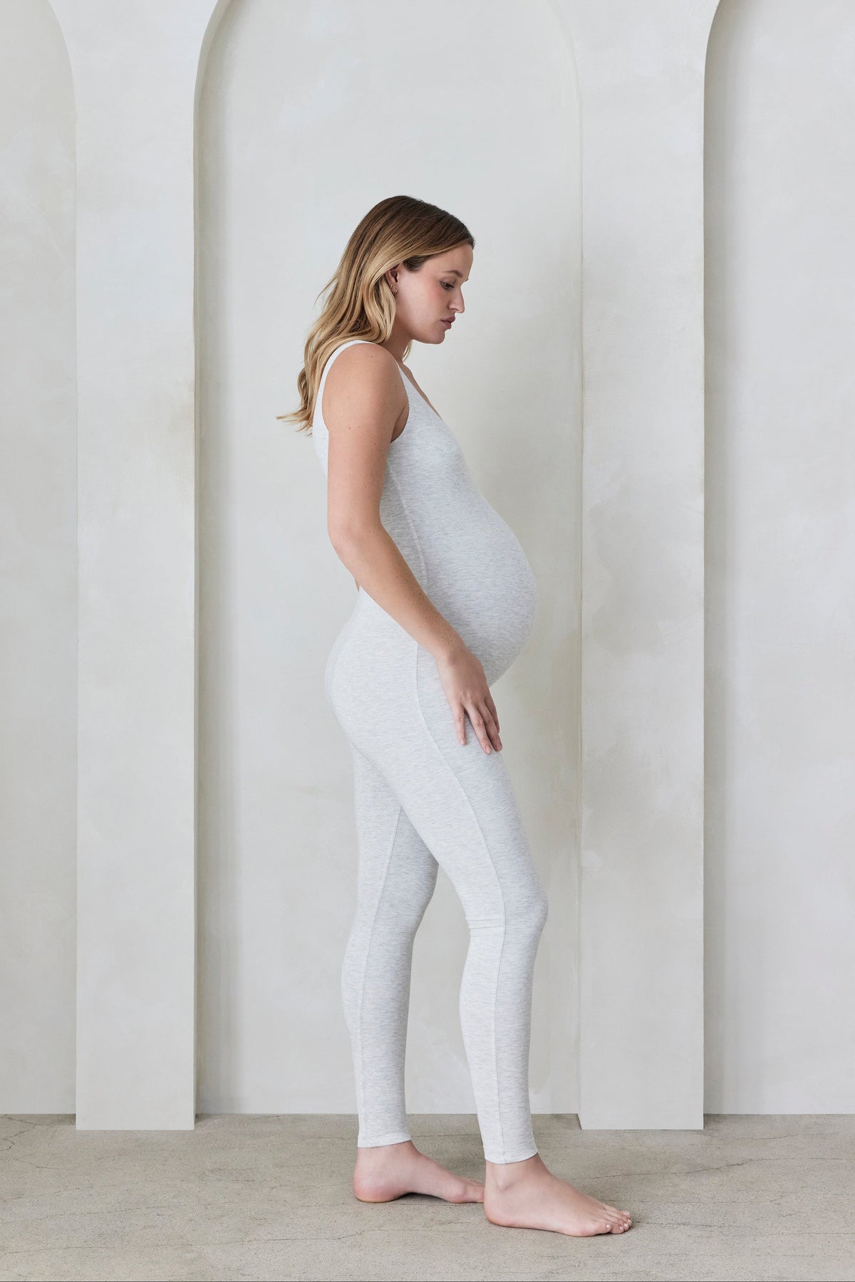 Pregnant woman wearing a heather grey Bumpsuit in a minimalistic setting with arches.