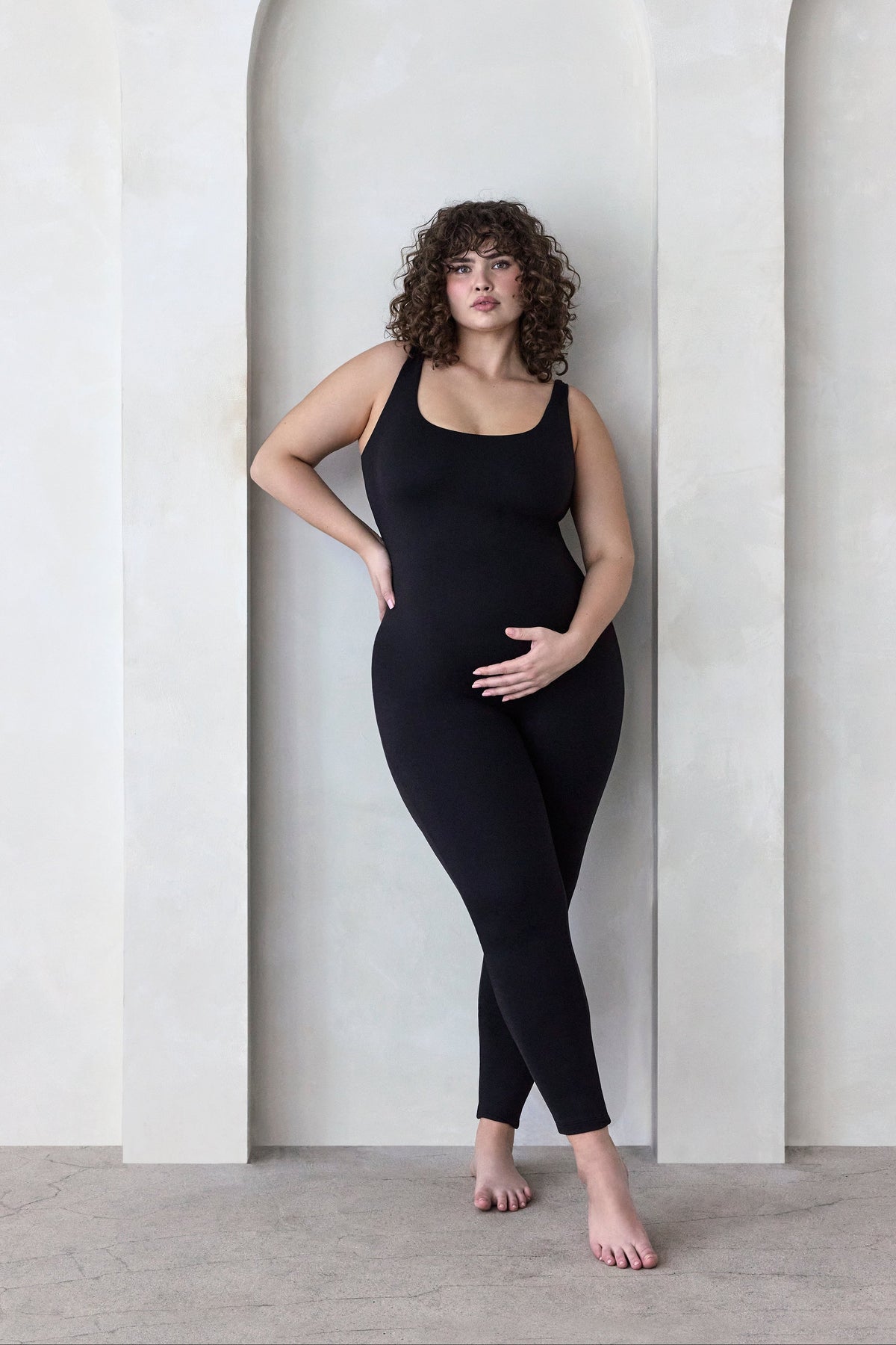 Woman wearing a black Bumpsuit Jumpsuit standing in a minimalistic setting with arches.