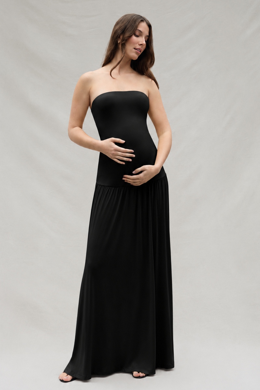 Woman holding her pregnant belly wearing a black Bumpsuit strapless maxi dress with a drop waist feature, standing in-front of an off white backdrop