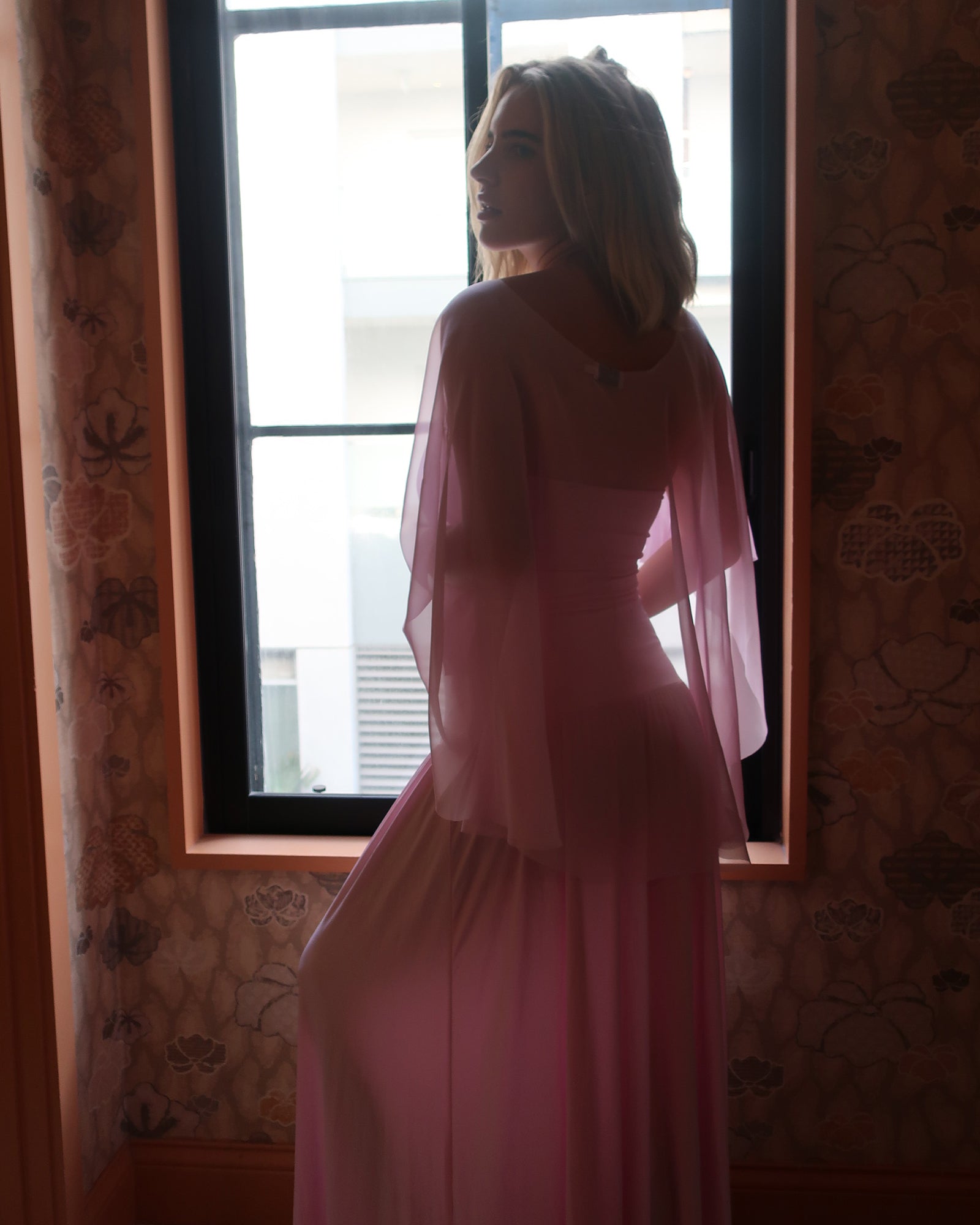 A pregnant woman stands in profile by a window wearing a flowing pink dress and chiffon cape by Bumpsuit , photographed in a dimly lit interior with patterned wallpaper.