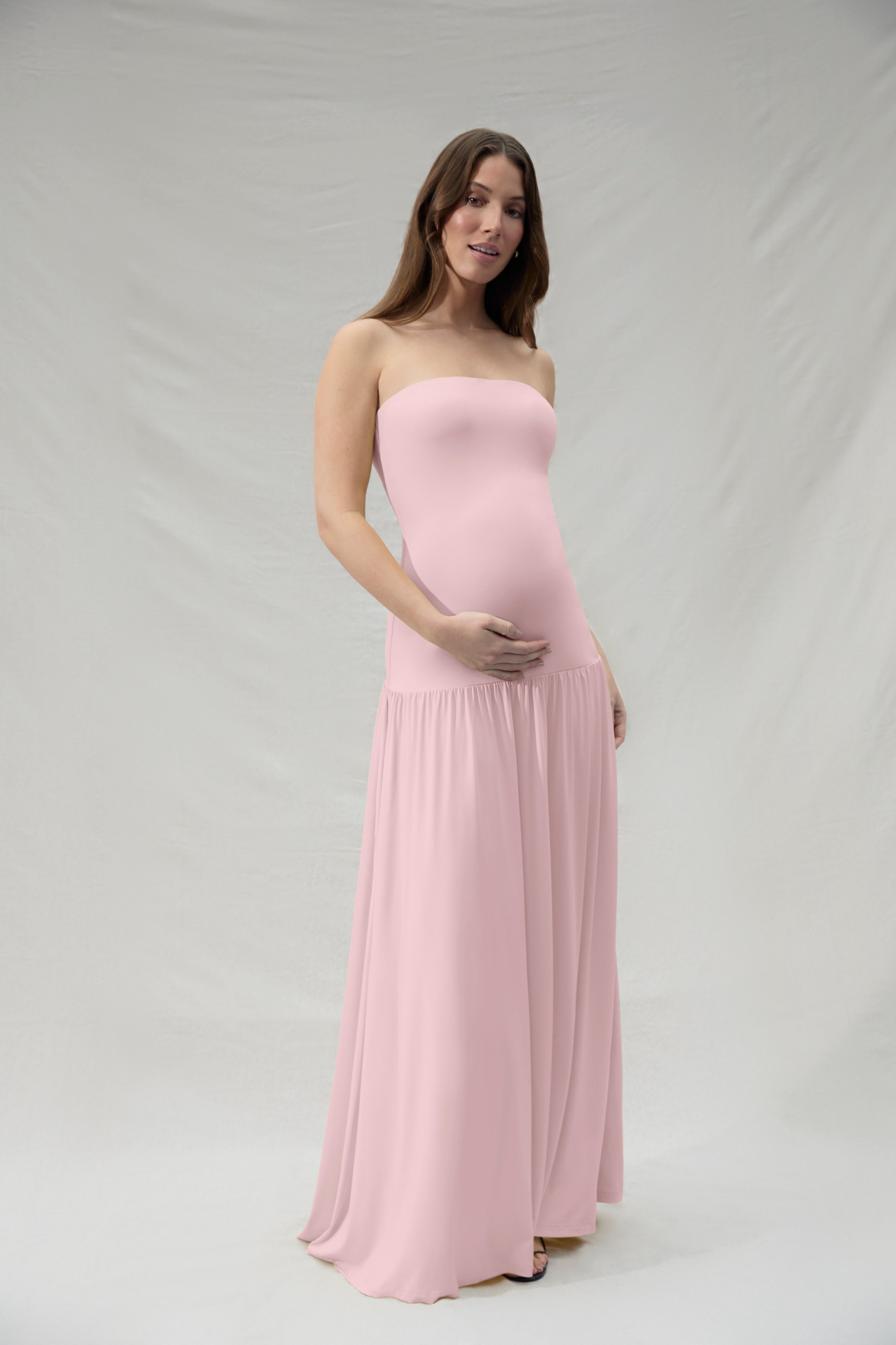 Pregnant woman wearing a Bumpsuit pink strapless matenrity gown against a plain background