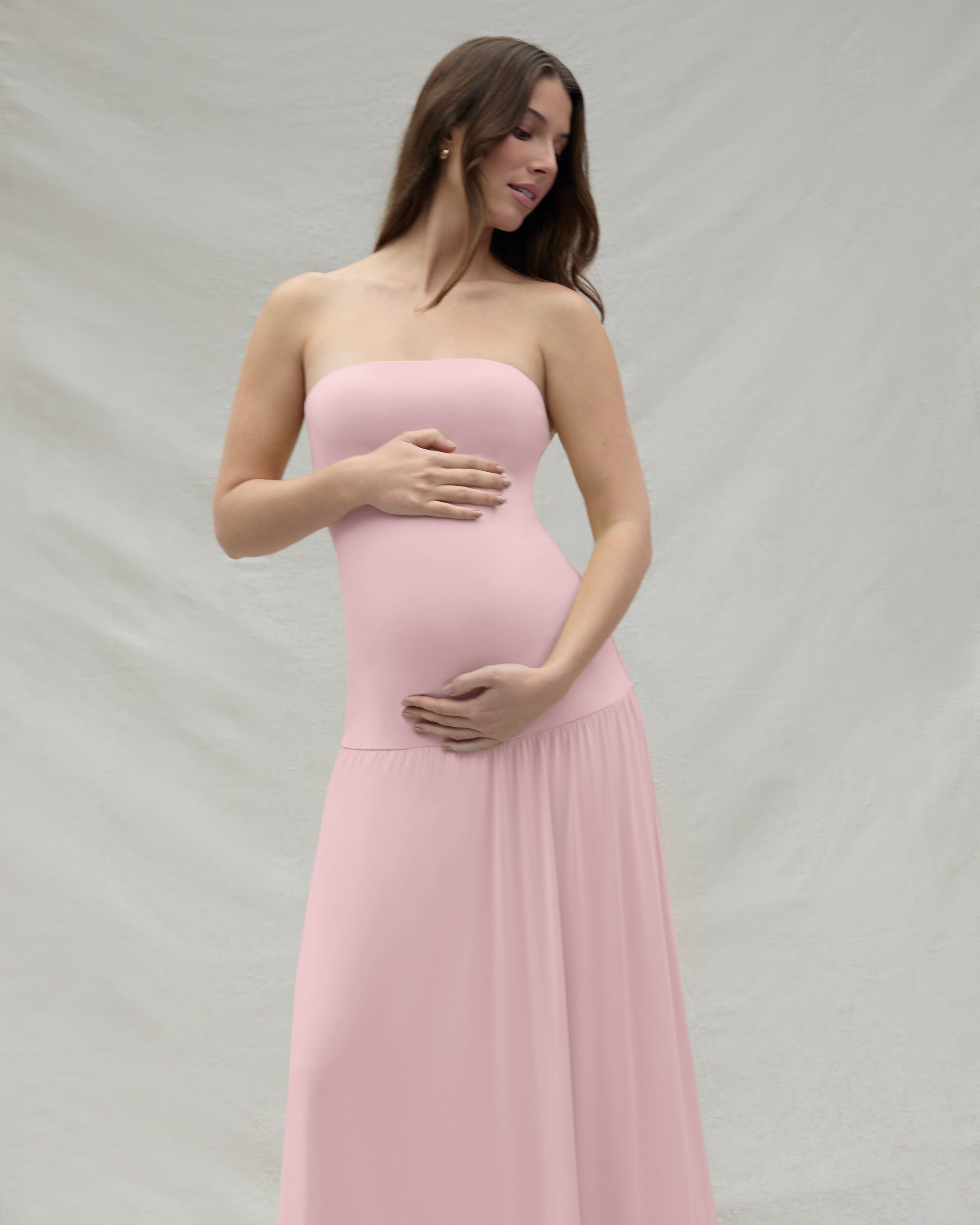 Pregnant woman wearing a pink strapless gown by Bumpsuit against a beige background