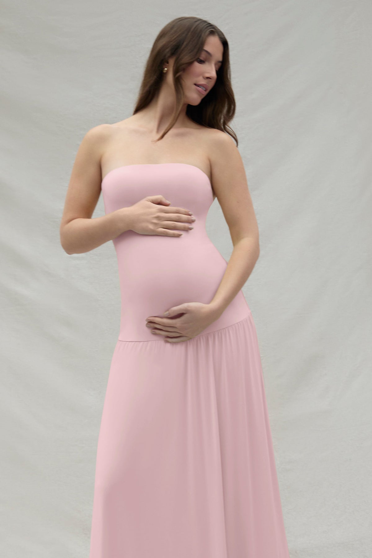 Pregnant woman wearing a pink strapless gown by Bumpsuit against a beige background