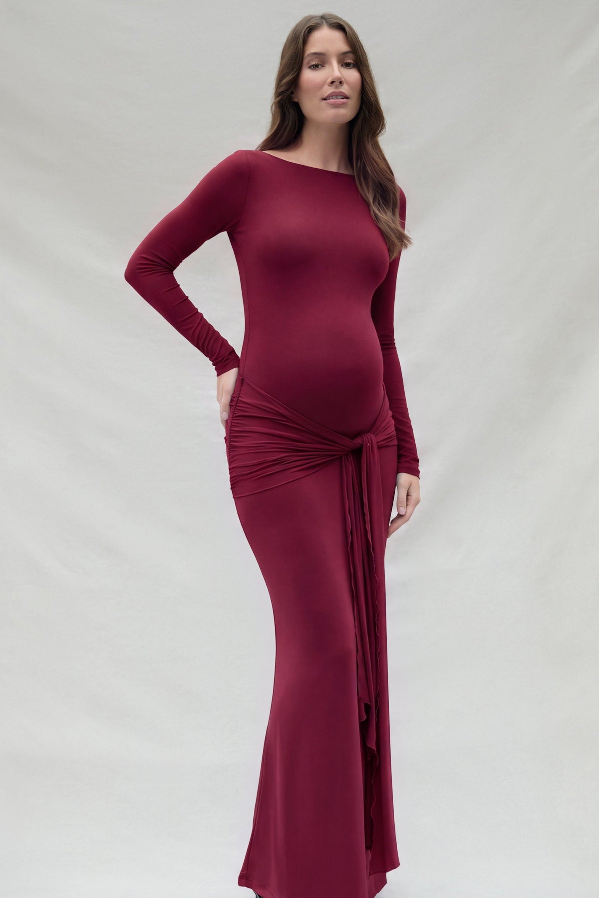 A pregnant woman wearing a long-sleeve, burgundy maxi Bumpsuit maternity dress with a draped wrap detail at the waist, standing against a light gray studio backdrop.