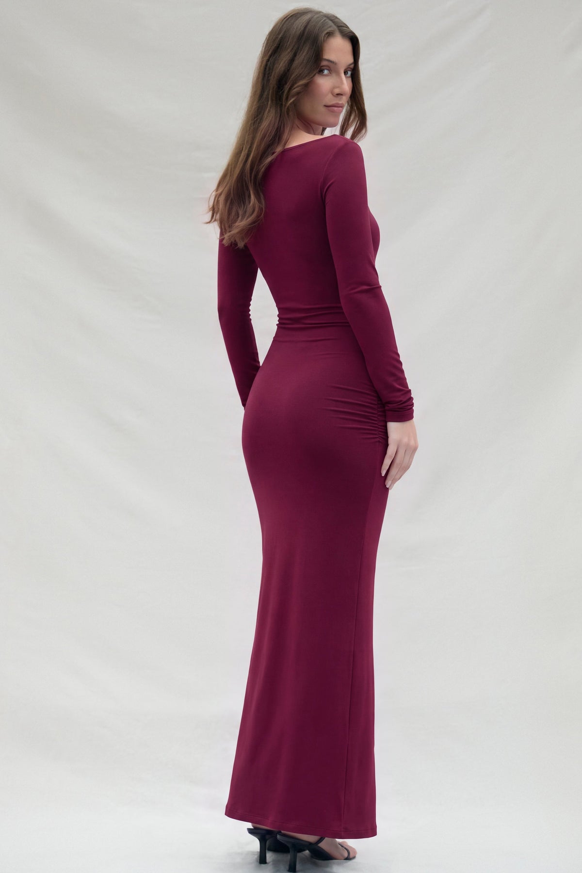 Back view of a woman modeling a long-sleeved, Bumpsuit maxi dress in burgundy,  against a beige studio backdrop.