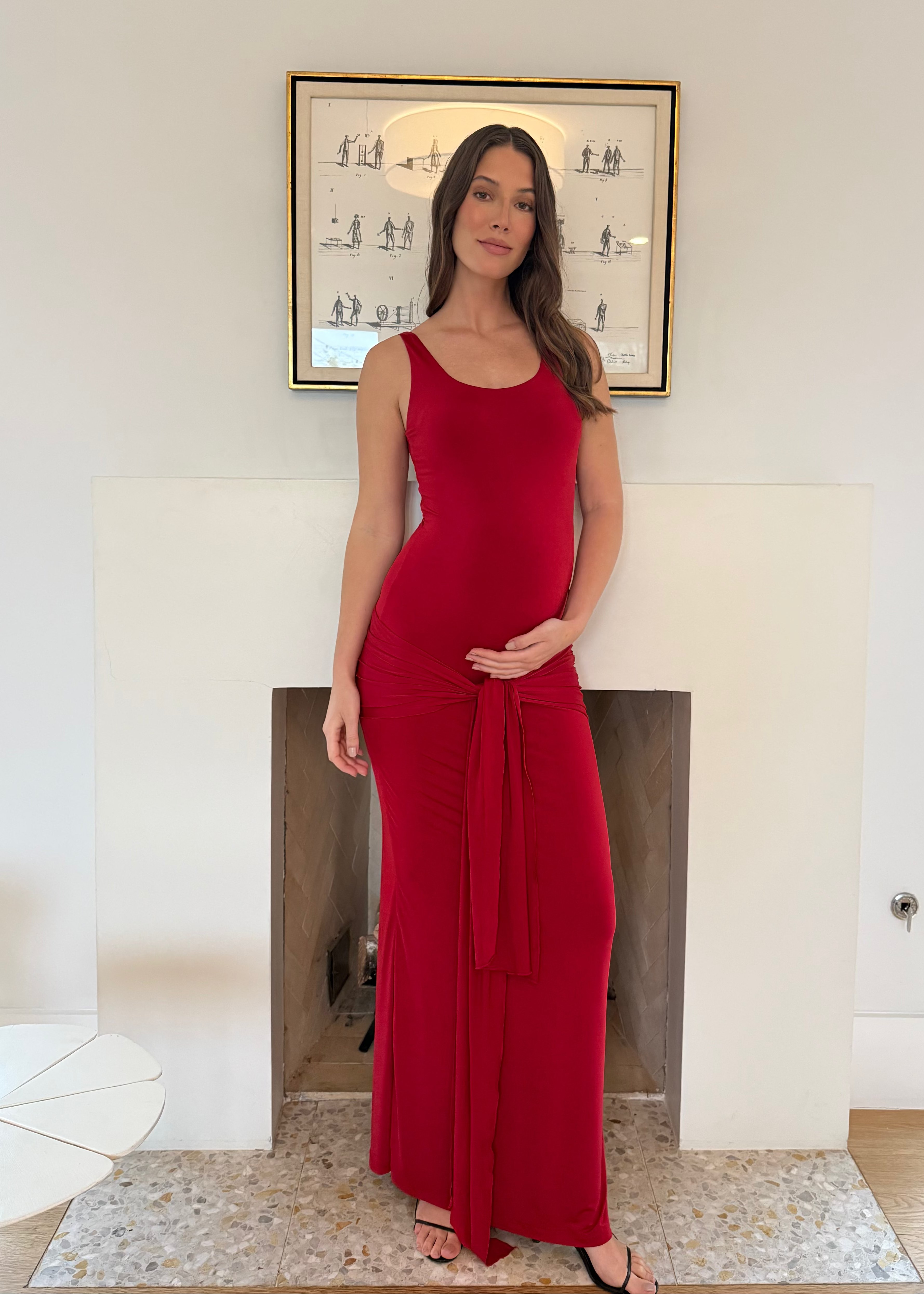 Pregnant woman in a Bumpsuit red maternity dress standing in a room with a fireplace and framed picture on the wall.