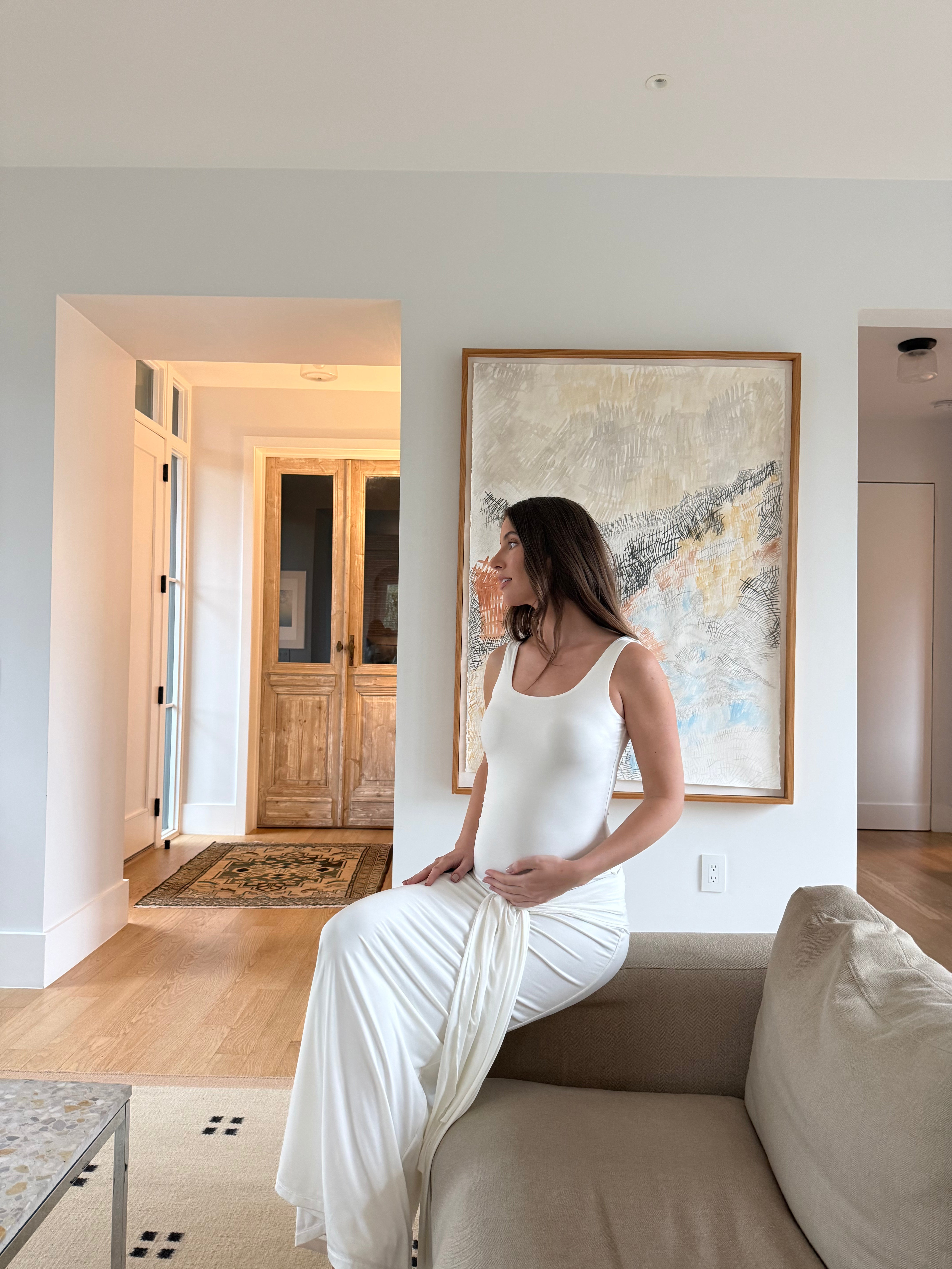 Pregnant woman in a white dress sitting on a couch in a modern living room with abstract art on the wall.