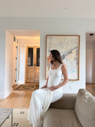 Pregnant woman in a white dress sitting on a couch in a modern living room with abstract art on the wall.