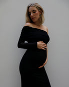 A pregnant woman wearing a Bumpsuit black off-the-shoulder long-sleeve maternity dress, posing with her hands on her belly against a plain gray wall.