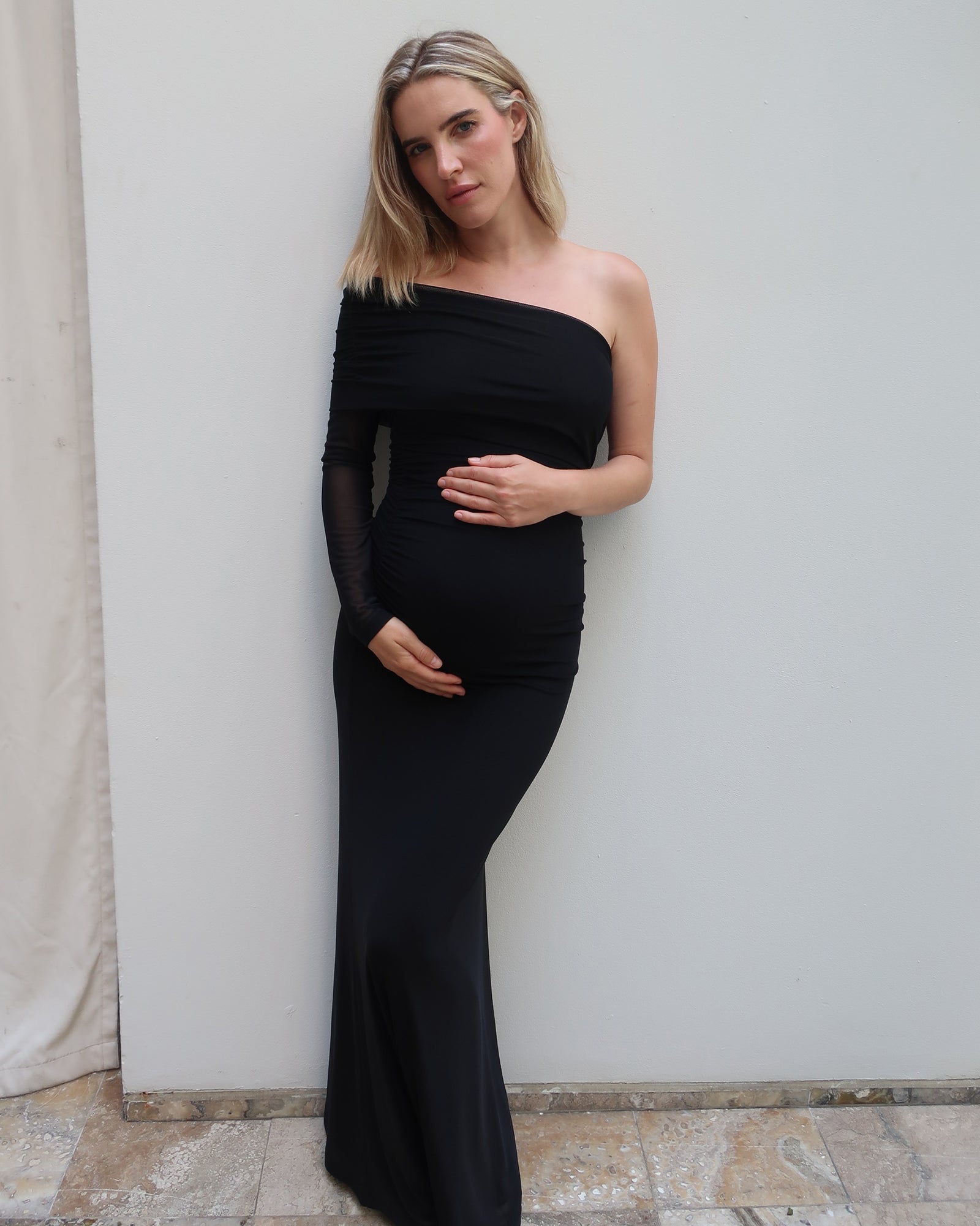 A pregnant woman with blonde hair models a black one-shoulder maternity gown with sheer long sleeves by Bumpsuit, posing against a plain light gray wall.