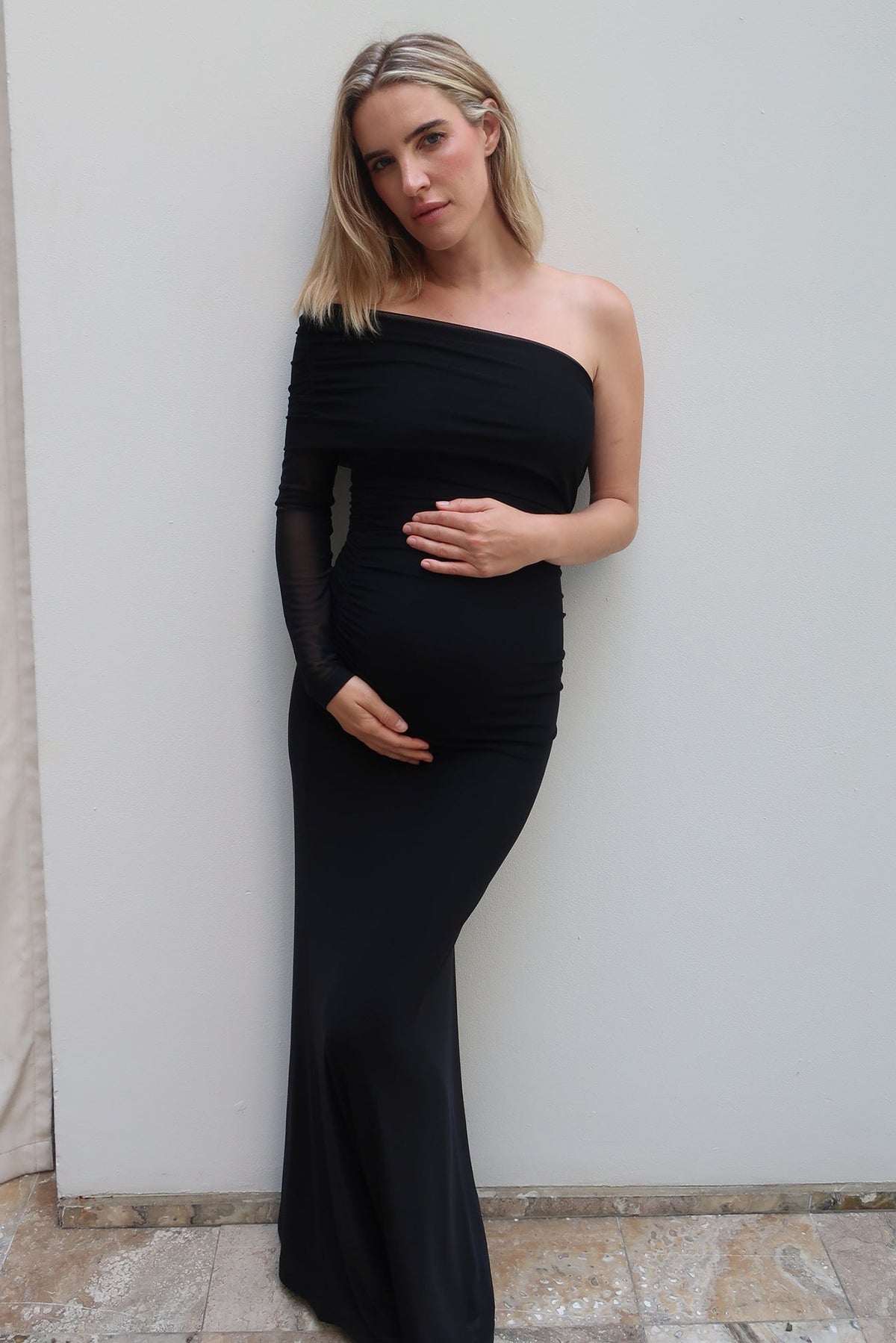 A pregnant woman with blonde hair models a black one-shoulder maternity gown with sheer long sleeves by Bumpsuit, posing against a plain light gray wall.