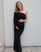A pregnant woman with blonde hair models a black one-shoulder maternity gown with sheer long sleeves by Bumpsuit, posing against a plain light gray wall.