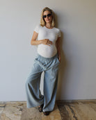 A pregnant woman wearing Bumpsuits light blue wide-leg denim pants and the everyday shirt, standing against a plain off-white wall on a stone tiled floor.