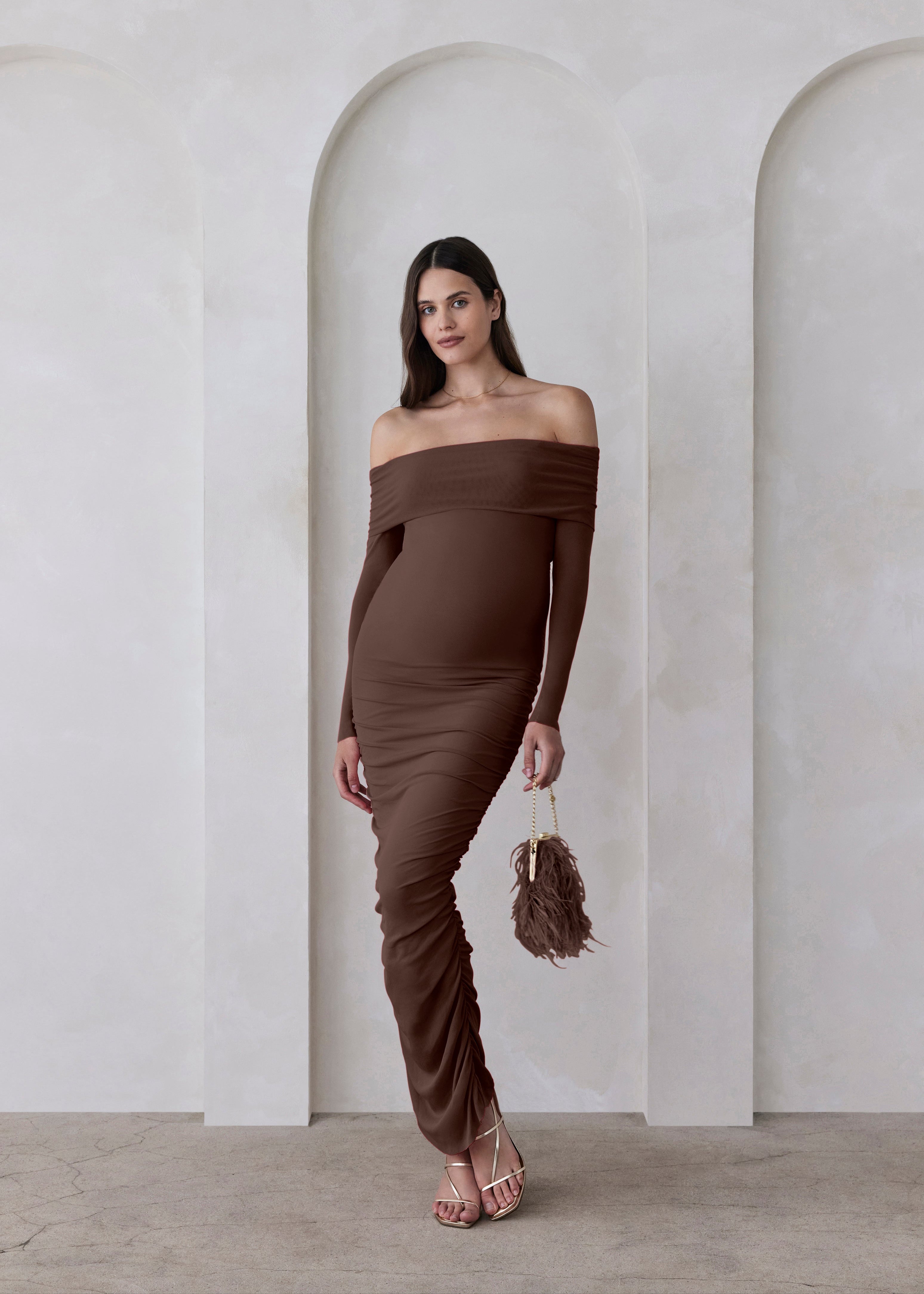 Woman wearing a brown off-shoulder dress standing in a minimalistic setting with arches.