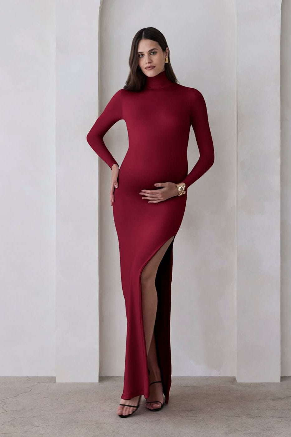 A pregnant woman wearing a burgundy turtleneck maternity maxi dress with long sleeves and a thigh-high slit on one side standing against a beige feature wall