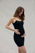 Woman wearing a black mini maternity slip  dress by Bumpsuit against a plain background