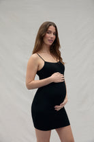 Woman holding her pregnant belly wearing a mini black dress against a plain background