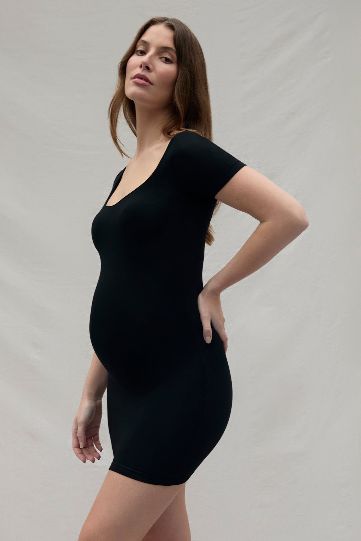 Pregnant woman wearing a black scoop neck cotton mini dress against a plain background