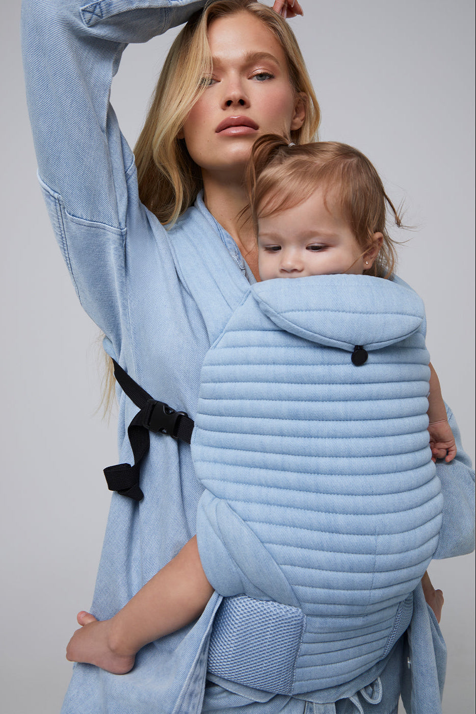 Front view of the light denim Armadillo baby carrier showcasing padded quilting and black straps on a model carrying a baby