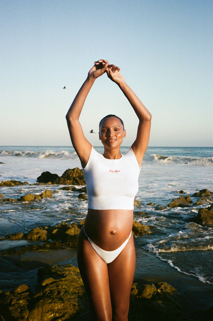 Woman in a white baby mama bumpsuit tee standing on a rocky beach with ocean waves in the background