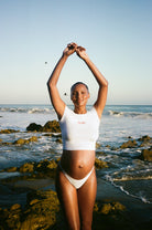Woman in a white baby mama bumpsuit tee standing on a rocky beach with ocean waves in the background
