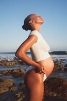 Pregnant woman standing on a rocky beach with ocean waves in the background wearing jumpsuits baby mama tee