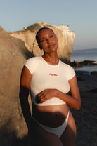 pregnant woman in a white bumpsuit outfit with 'Baby mama' wording standing on a beach with cliffs and ocean in the background.