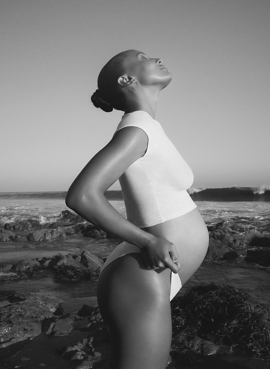Black and white photo of a pregnant woman standing on a rocky beach.