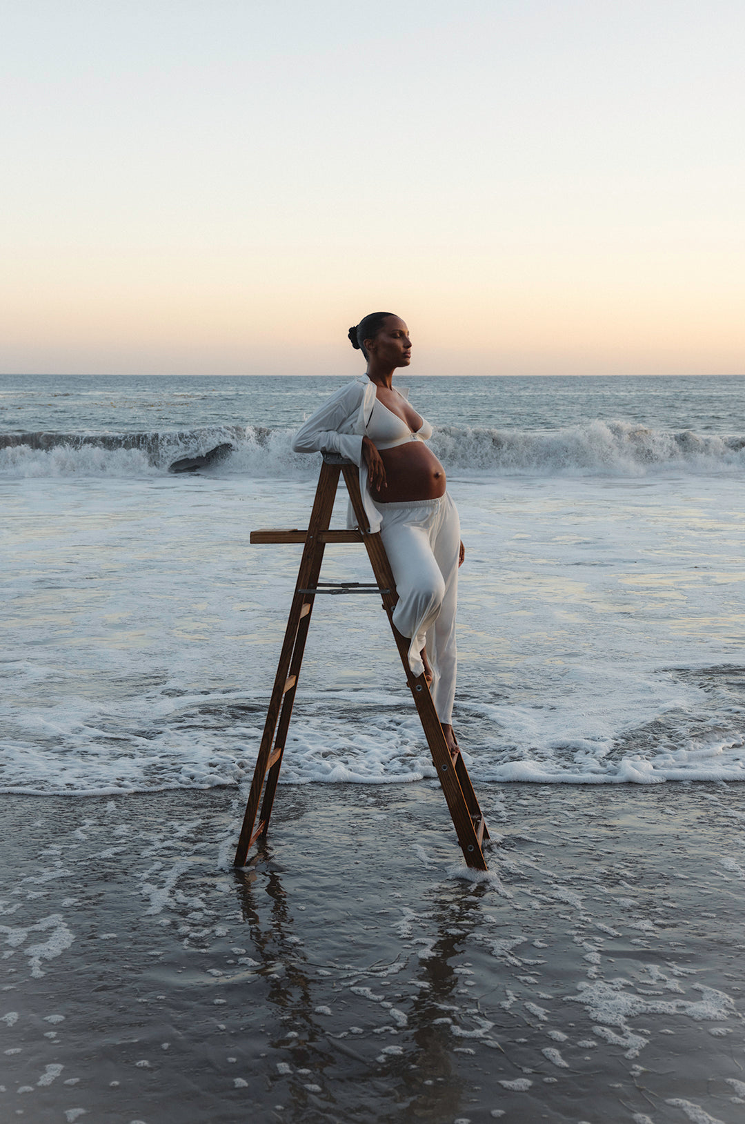 Jasmine tookes standing on a wooden ladder in the ocean with a sunset background wearing Bumpsuit Maternity Luxe Bamboo pjs in white