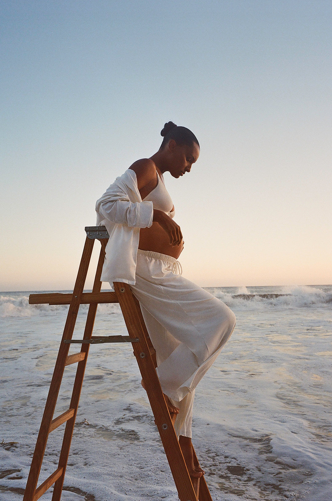 Pregnant model sitting on a ladder in the ocean wearing bumpsuit white Luxe Pjs in bamboo