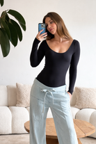 Pregnant woman taking a mirror selfie wearing bumpsuits black long-sleeve scoop neck bodysuit and light blue pants in a living room.