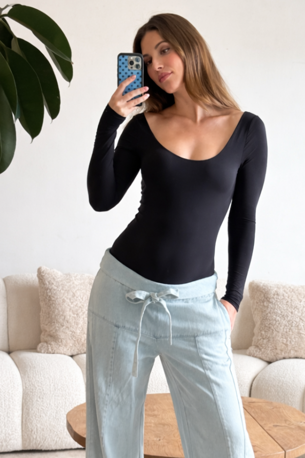 Pregnant woman taking a mirror selfie wearing bumpsuits black long-sleeve scoop neck bodysuit and light blue pants in a living room.