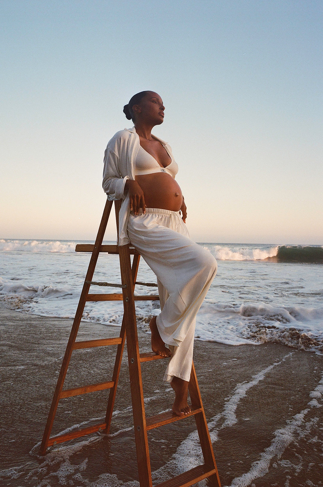 Jasmine Tookes standing on a ladder by the ocean at sunset wearing bumpsuits Luxe Bamboo Pj set