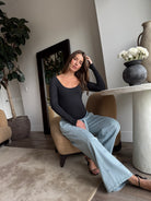 Pregnant woman sitting in a chair in a modern living room wearing Bumpsuits long sleeve scoop neck bodysuit and jeans 
