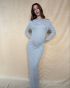 A woman wearing a heather grey long sleeve maternity dress, against  a beige fabric surface.