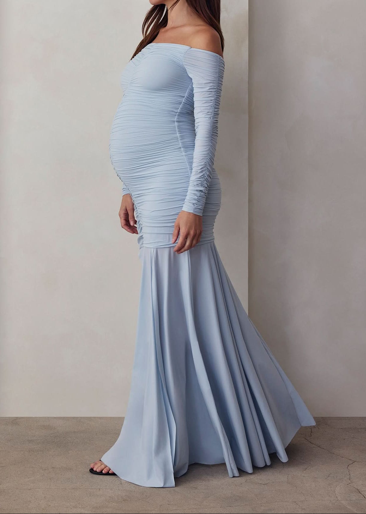 A pregnant woman wearing an off-the-shoulder, maxi-length, blue maternity dress with a flowing skirt.
