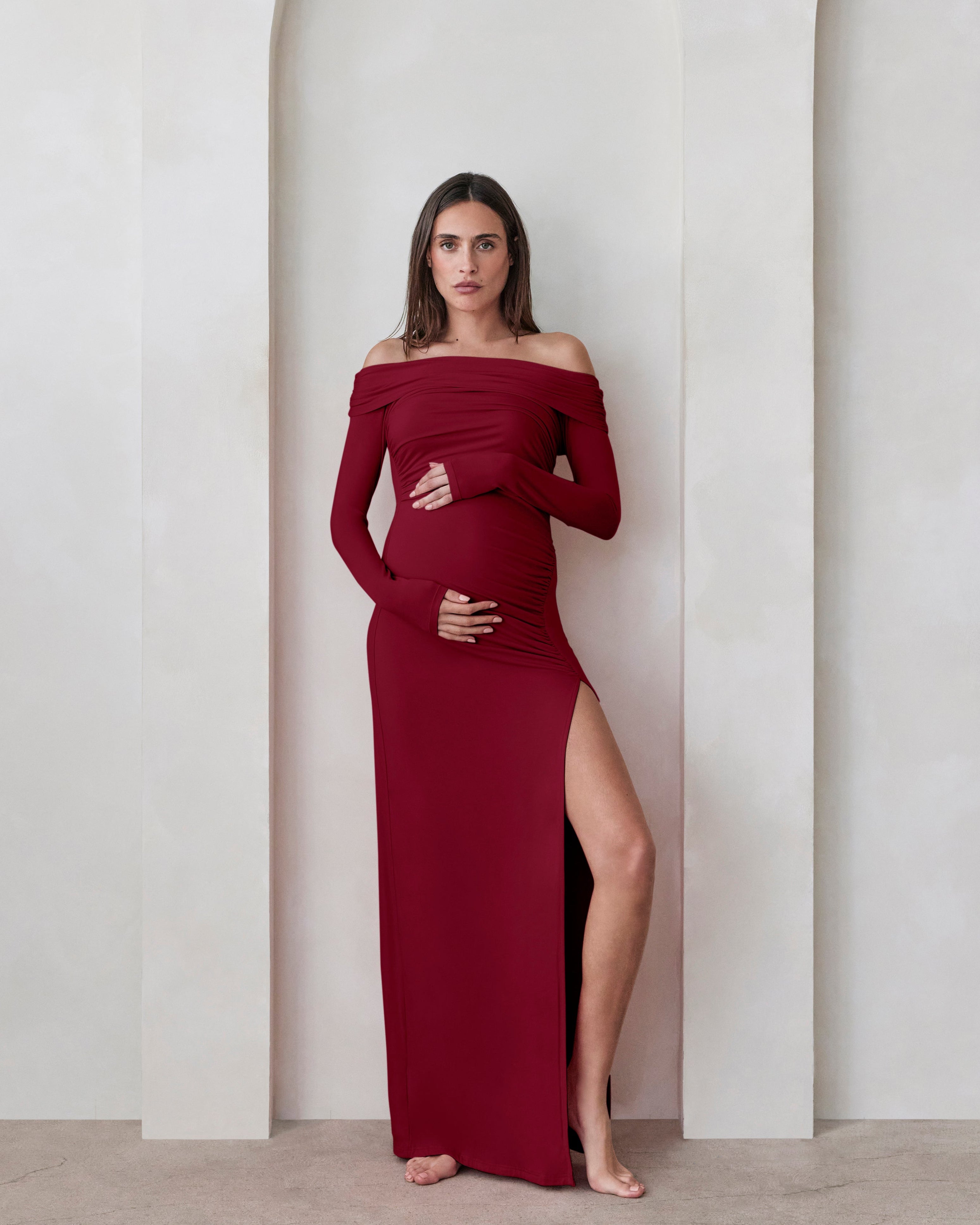 A pregnant woman wearing a Bumpsuit burgundy off the shoulder maxi dress with  high leg slit against a beige feature wall
