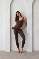 A woman modeling a matching chocolate brown tank top and leggings set by Bumpsuit, standing barefoot in front of a white wall with three arched recesses.