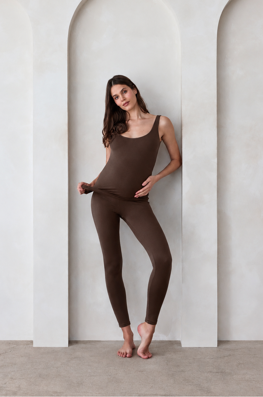 A woman modeling a matching chocolate brown tank top and leggings set by Bumpsuit, standing barefoot in front of a white wall with three arched recesses.