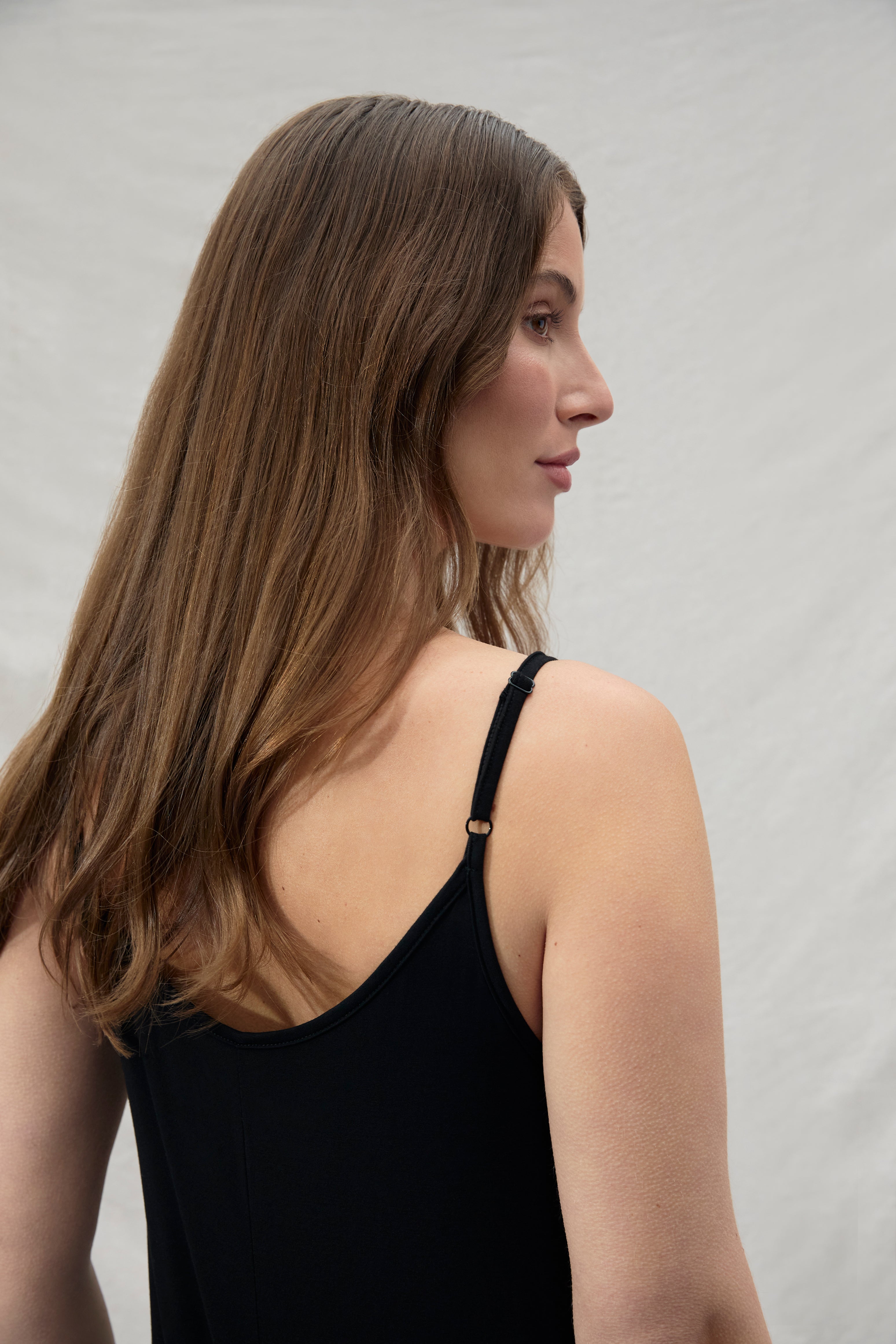 back side view of a woman wearing a black sleeveless jumpsuit looking away against a plain background