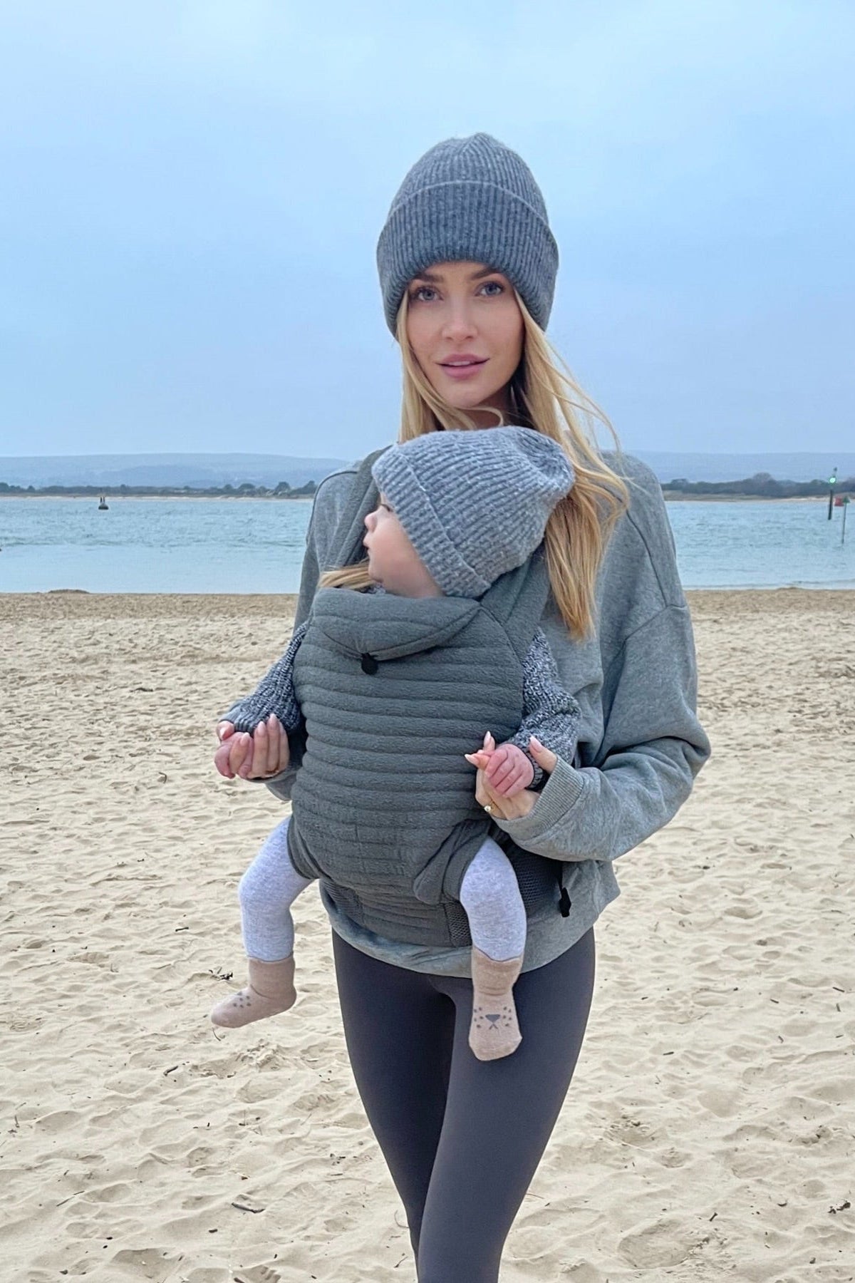 woman on the beach with a baby wearing the slate coloured armadillo baby carrier
