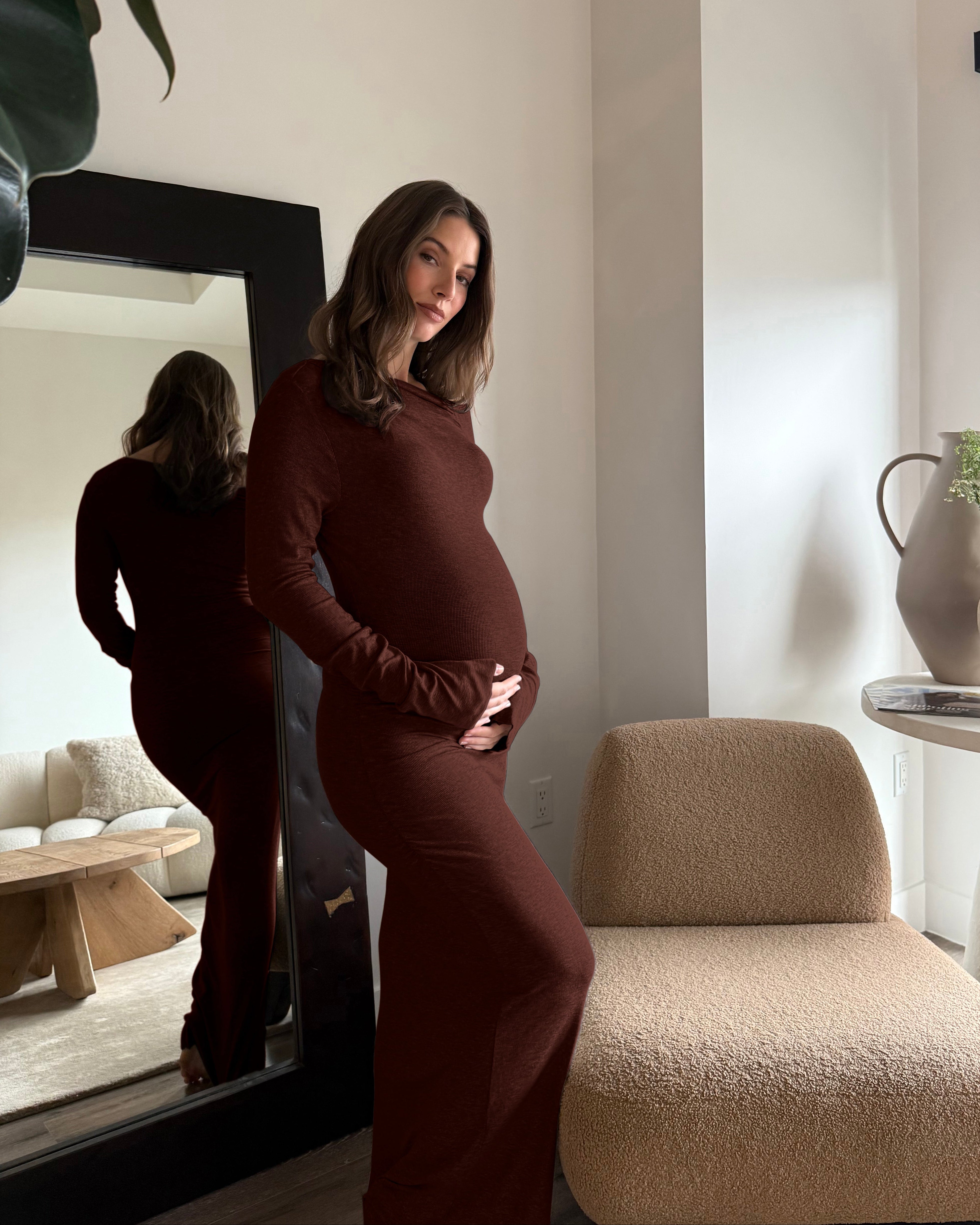 A pregnant woman wearing a chocolate brown long sleeve soft rib maxi dress in a modern room, with her reflection visible in a large black-framed mirror.