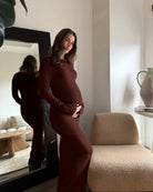 A pregnant woman wearing a chocolate brown long sleeve soft rib maxi dress in a modern room, with her reflection visible in a large black-framed mirror.