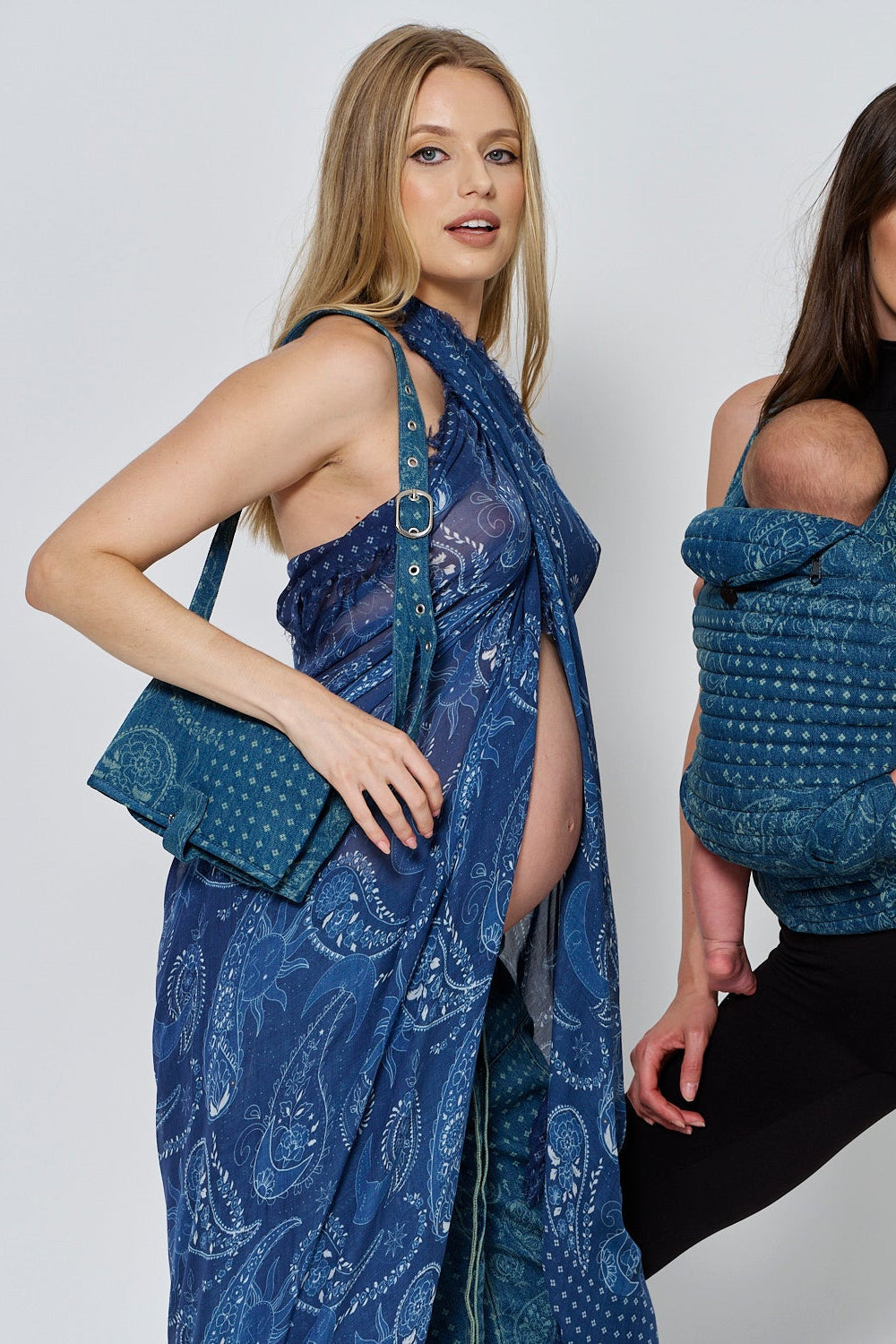 Two women model blue patterned Spell and BUMPSUIT accessories, including a shoulder bag and baby carrier