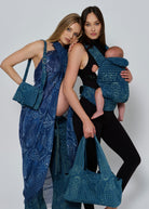 Two women holding blue spell and bumpsuit  textured bags with a baby in a carrier against a plain background